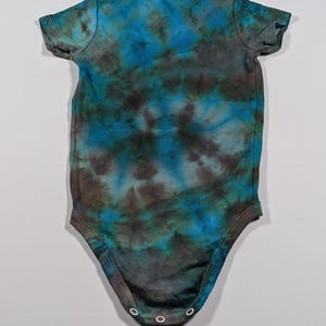 May include: A tie-dye baby bodysuit in shades of blue, teal, and brown. The short-sleeved onesie has a round neckline and three snap closures at the bottom. The fabric has a mottled, watercolor-like appearance.