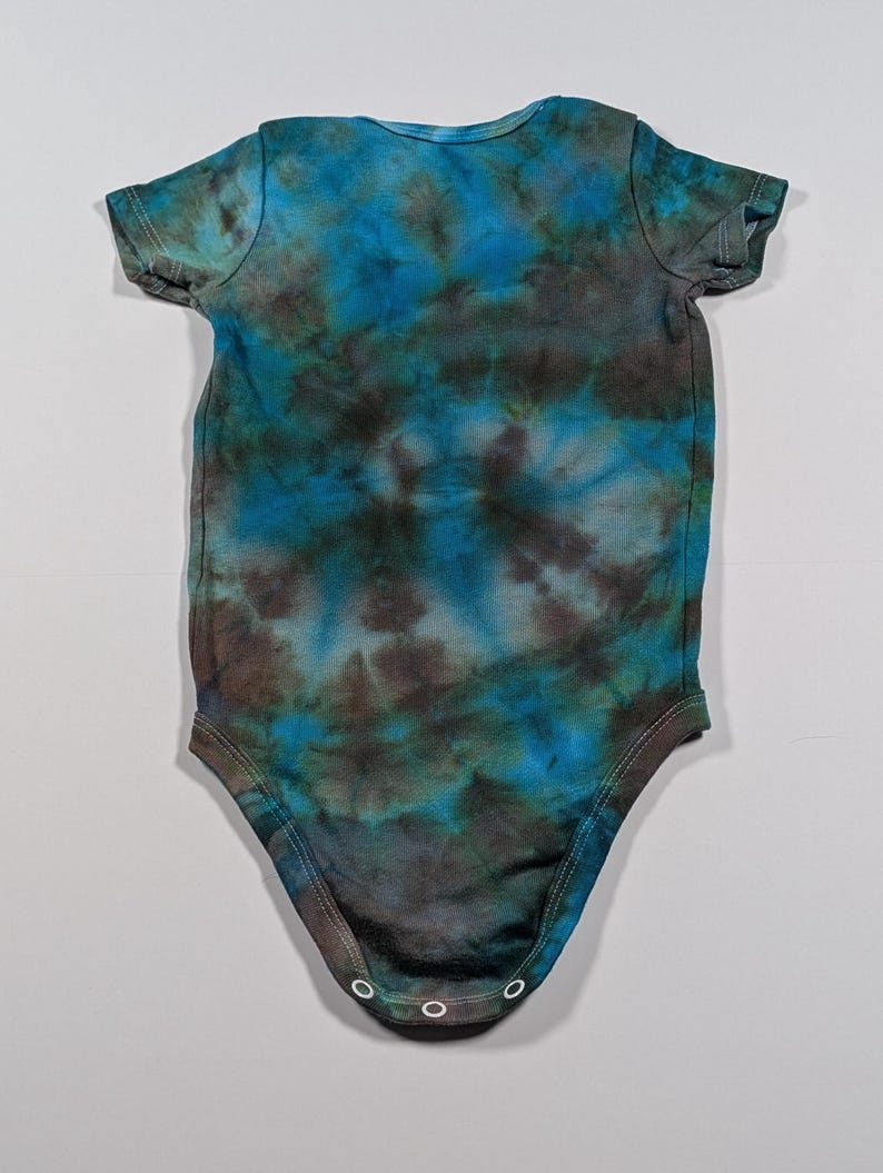 May include: A tie-dye baby bodysuit in shades of blue, teal, and brown. The short-sleeved onesie has a round neckline and three snap closures at the bottom. The fabric has a mottled, watercolor-like appearance.