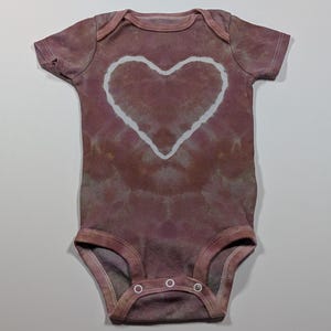 May include: A tie-dye baby onesie in shades of mauve and brown, featuring a large white heart design in the center. The onesie has short sleeves and snap closures at the bottom, perfect for a baby.