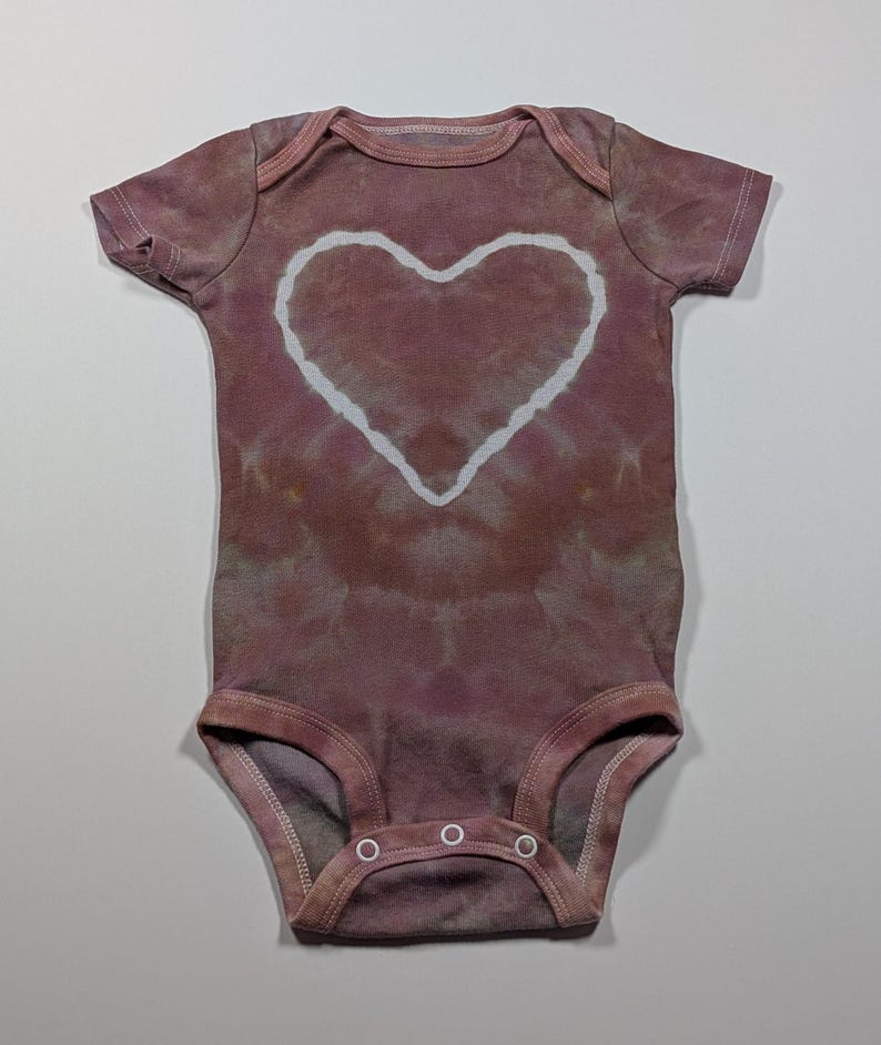 May include: A tie-dye baby onesie in shades of mauve and brown, featuring a large white heart design in the center. The onesie has short sleeves and snap closures at the bottom, perfect for a baby.
