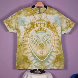 May include: A tie-dye t-shirt with an alien face design. The shirt features a blend of yellow, blue, and white colors. The alien face is centered on the front of the shirt, with large eyes and a simple outline. The shirt is hanging on a wooden hanger.