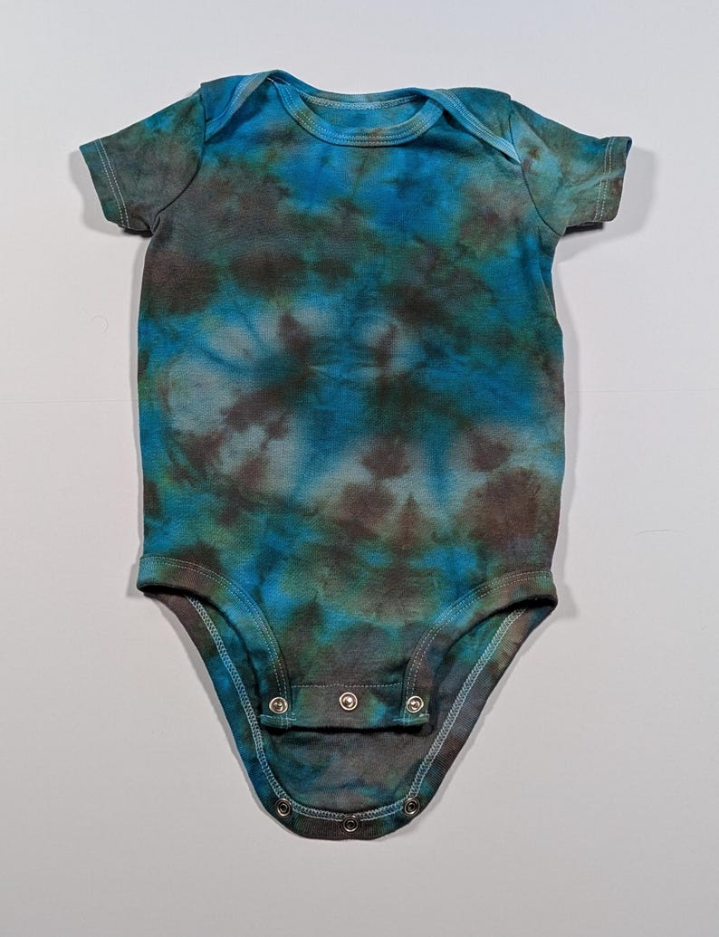May include: A short-sleeved baby bodysuit with a tie-dye pattern in shades of blue, teal, and brown. The bodysuit has a round neckline and snaps at the bottom for easy changes. The fabric appears to be soft and comfortable.