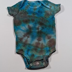 May include: A tie-dye baby onesie in shades of blue, teal, and brown. The short-sleeved garment has a round neckline and three snap closures at the bottom. The fabric has a mottled, swirled pattern.