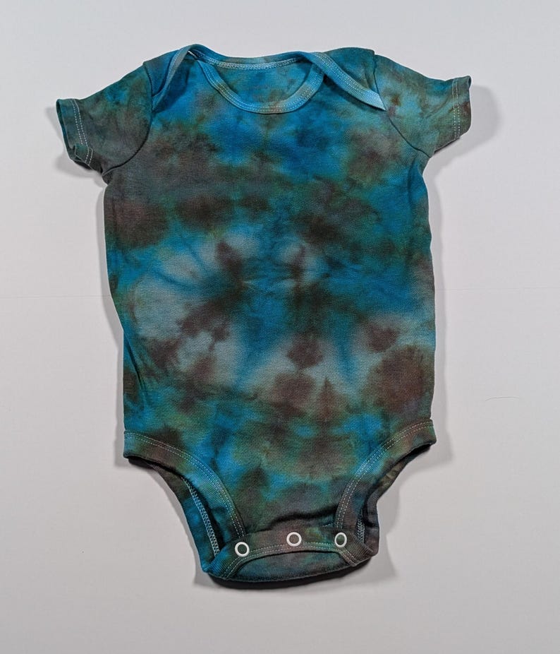 May include: A tie-dye baby onesie in shades of blue, teal, and brown. The short-sleeved garment has a round neckline and three snap closures at the bottom. The fabric has a mottled, swirled pattern.
