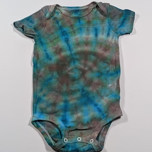 Puede incluir: Un body de beb&eacute; te&ntilde;ido con tinte anudado en tonos azules, verdes y marrones. La prenda de manga corta tiene un cuello redondo y tres cierres de presi&oacute;n en la parte inferior. La tela tiene un patr&oacute;n moteado.