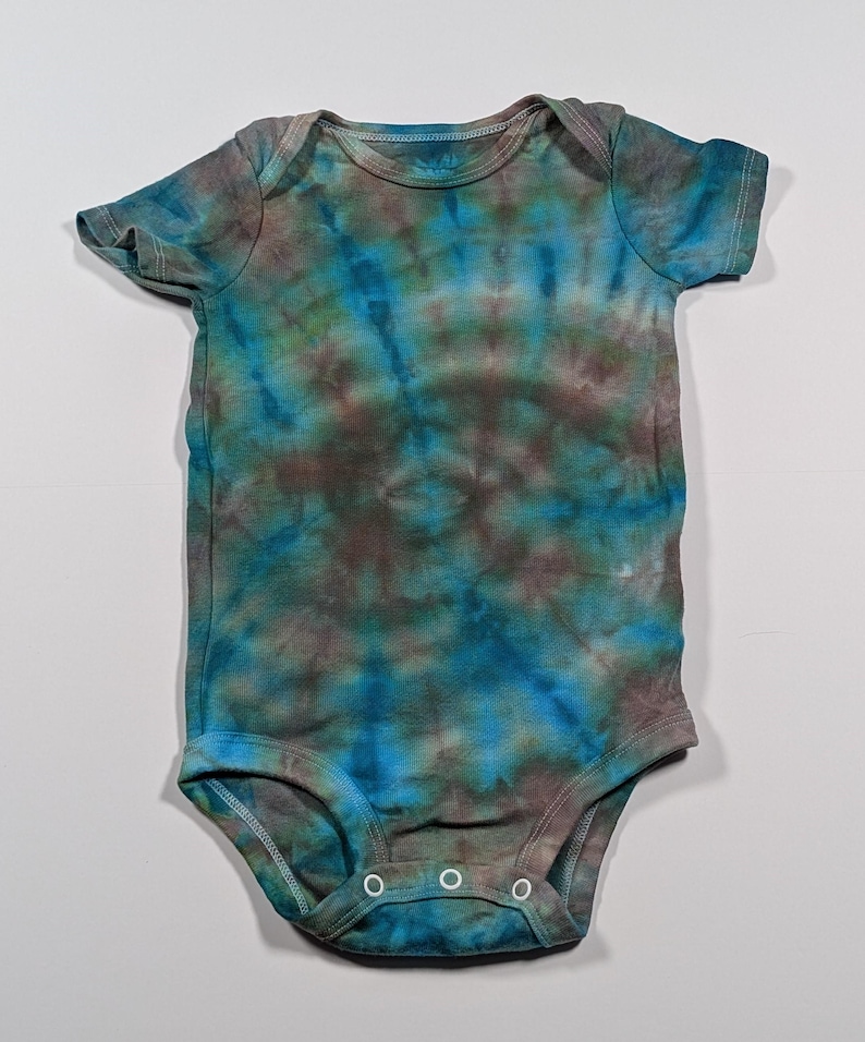 Puede incluir: Un body de beb&eacute; te&ntilde;ido con tinte anudado en tonos azules, verdes y marrones. La prenda de manga corta tiene un cuello redondo y tres cierres de presi&oacute;n en la parte inferior. La tela tiene un patr&oacute;n moteado.
