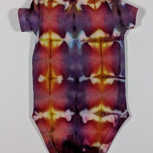 Puede incluir: Un body de beb&eacute; te&ntilde;ido anudado en tonos morados, rojos y amarillos. El body de manga corta presenta un patr&oacute;n sim&eacute;trico con corchetes blancos en la parte inferior. La tela tiene un aspecto suave y texturizado.