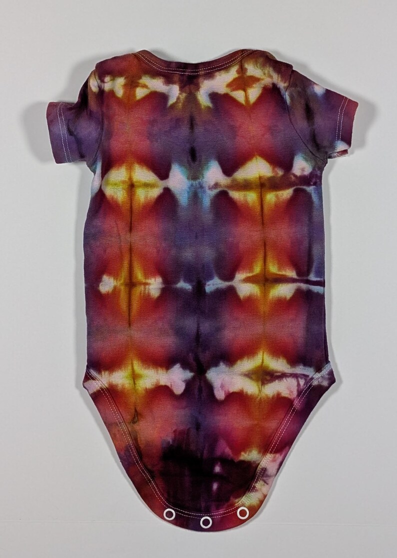 Puede incluir: Un body de beb&eacute; te&ntilde;ido anudado en tonos morados, rojos y amarillos. El body de manga corta presenta un patr&oacute;n sim&eacute;trico con corchetes blancos en la parte inferior. La tela tiene un aspecto suave y texturizado.