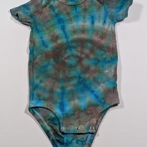 Puede incluir: Un body de beb&eacute; te&ntilde;ido anudado en tonos azules, marrones y verdes. El body de manga corta tiene un cuello redondo y corchetes en la parte inferior. La tela tiene un patr&oacute;n moteado y arremolinado.