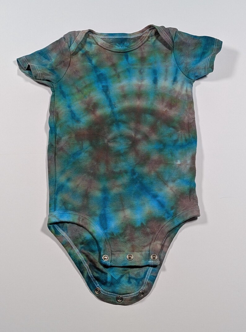 Puede incluir: Un body de beb&eacute; te&ntilde;ido anudado en tonos azules, marrones y verdes. El body de manga corta tiene un cuello redondo y corchetes en la parte inferior. La tela tiene un patr&oacute;n moteado y arremolinado.