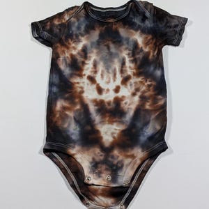 May include: A tie-dye baby bodysuit in shades of brown, black, and white. The short-sleeved onesie features a round neckline and snap closures at the bottom. The brand name "Carter's" is visible on the neckline.