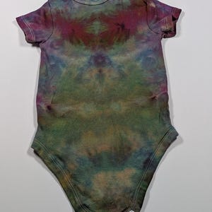 May include: A tie-dye baby onesie in shades of green, purple, and blue. The short-sleeved garment has a round neckline and snaps at the bottom. The fabric has a mottled, watercolor-like appearance.