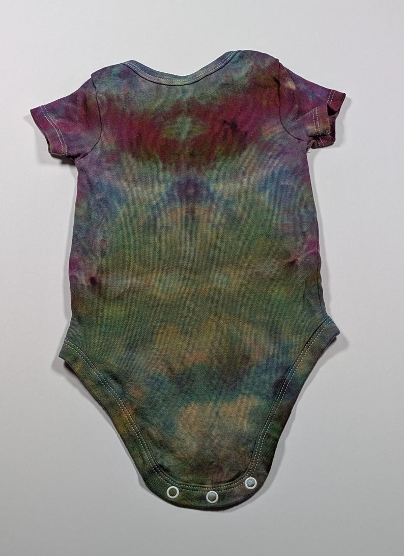 May include: A tie-dye baby onesie in shades of green, purple, and blue. The short-sleeved garment has a round neckline and snaps at the bottom. The fabric has a mottled, watercolor-like appearance.