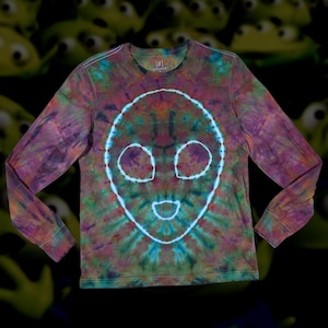 May include: A long-sleeved tie-dye shirt featuring a white alien face design on the front. The shirt's colors include shades of purple, green, and brown. The alien face has large eyes and a smiling mouth. The shirt is a casual wear item.