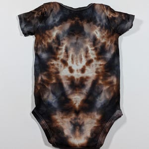 May include: A tie-dye baby onesie in shades of brown, black, and white. The short-sleeved garment has a symmetrical pattern with white stitching around the edges. The onesie is laid flat on a white surface.