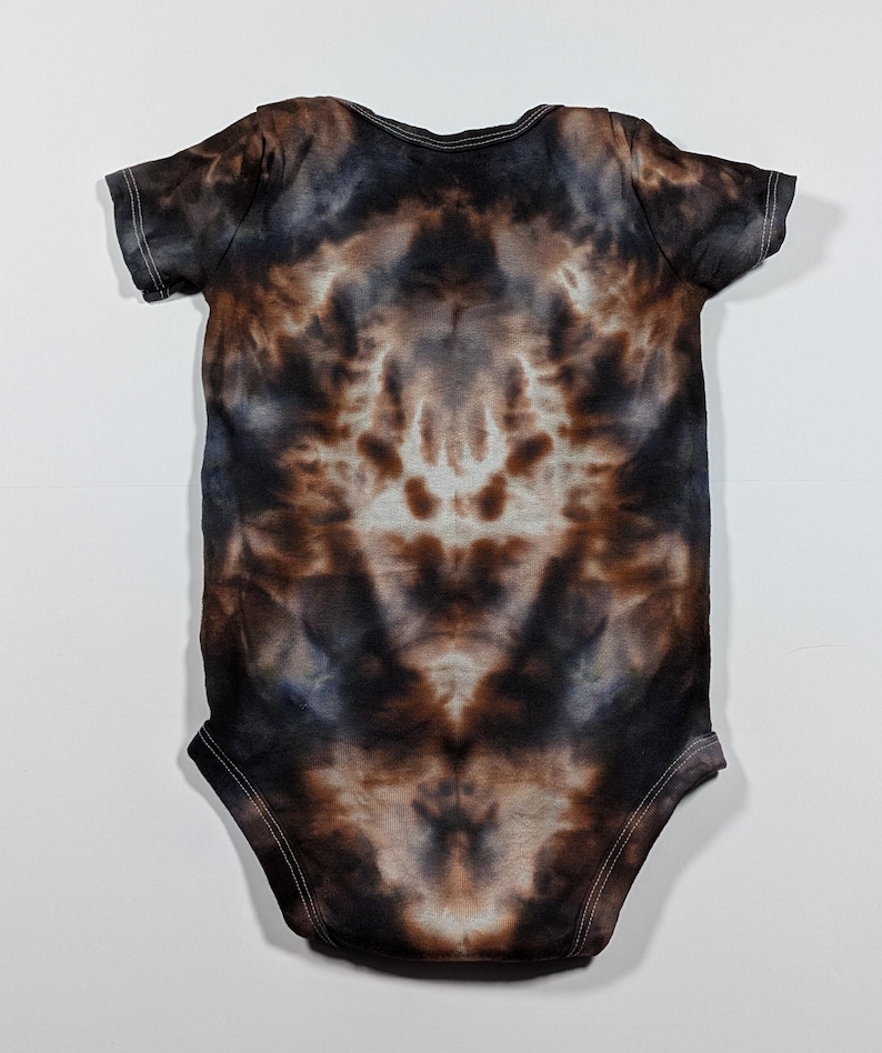 May include: A tie-dye baby onesie in shades of brown, black, and white. The short-sleeved garment has a symmetrical pattern with white stitching around the edges. The onesie is laid flat on a white surface.
