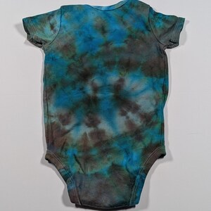 May include: A tie-dye baby onesie in shades of blue, teal, and brown. The short-sleeved bodysuit has a round neckline and snaps at the bottom. The fabric has a mottled, cloud-like pattern.