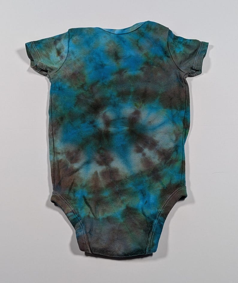 May include: A tie-dye baby onesie in shades of blue, teal, and brown. The short-sleeved bodysuit has a round neckline and snaps at the bottom. The fabric has a mottled, cloud-like pattern.