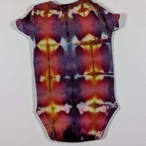 Puede incluir: Un body de beb&eacute; te&ntilde;ido con tinte anudado en tonos morado, rojo y amarillo. El body de manga corta presenta un patr&oacute;n sim&eacute;trico, cuello redondo y cierres de presi&oacute;n. La tela es suave, probablemente de algod&oacute;n. Para beb&eacute;s.