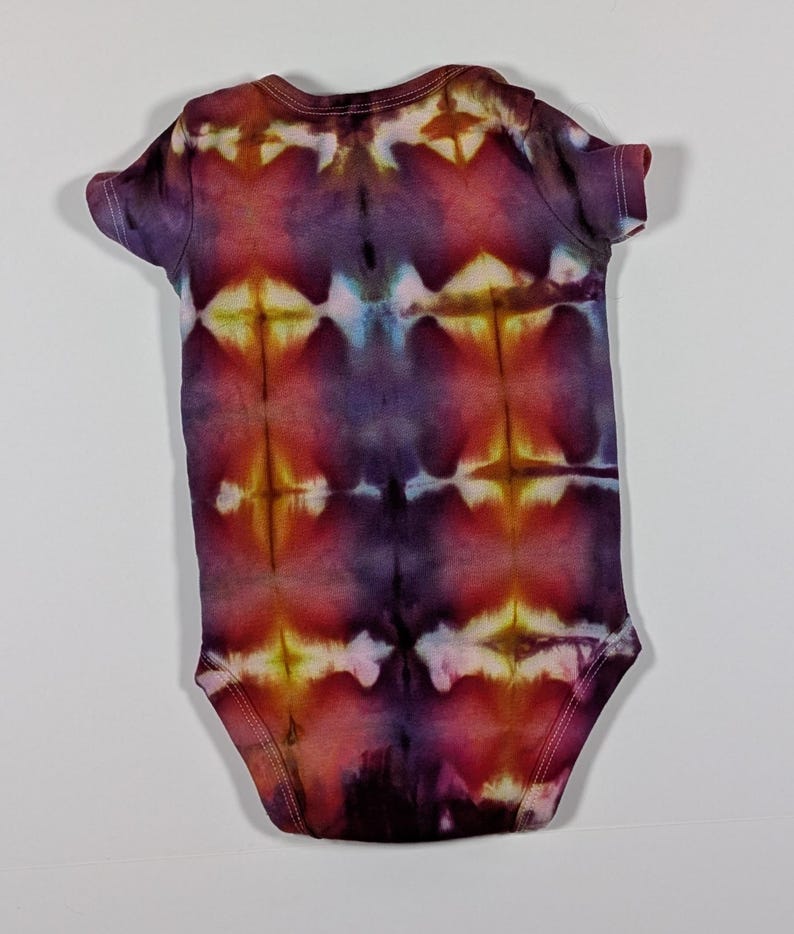 Puede incluir: Un body de beb&eacute; te&ntilde;ido con tinte anudado en tonos morado, rojo y amarillo. El body de manga corta presenta un patr&oacute;n sim&eacute;trico, cuello redondo y cierres de presi&oacute;n. La tela es suave, probablemente de algod&oacute;n. Para beb&eacute;s.