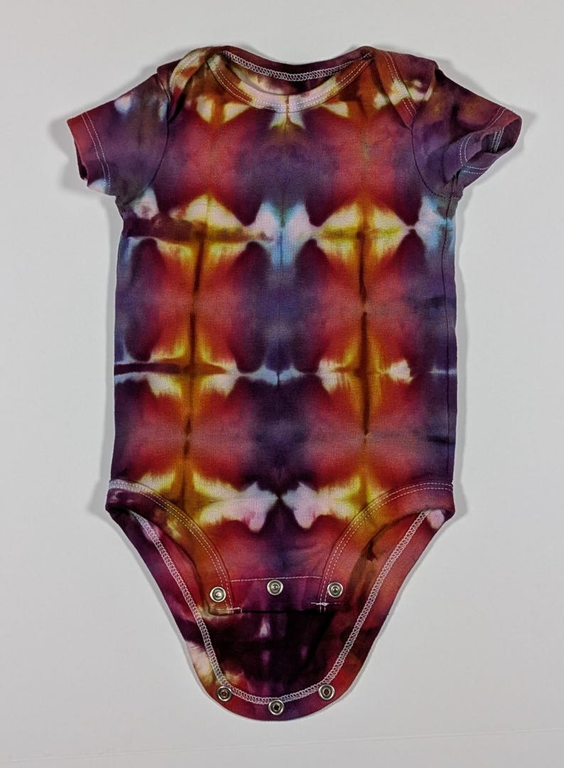 Puede incluir: Un body de beb&eacute; te&ntilde;ido con tinte en tonos morado, rojo y amarillo. El body de manga corta tiene un cierre de broches en la parte inferior y un cuello redondo. El patr&oacute;n colorido es sim&eacute;trico, con un dise&ntilde;o &uacute;nico.