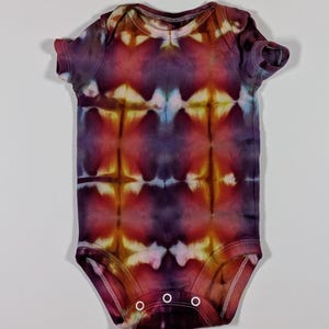 Puede incluir: Un body de beb&eacute; te&ntilde;ido con tinte en tonos morado, rojo y amarillo. La prenda de manga corta tiene un cuello redondo y tres cierres de bot&oacute;n a presi&oacute;n en la parte inferior. El patr&oacute;n tie-dye es sim&eacute;trico, con una mezcla de colores.