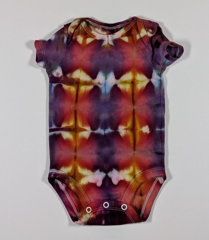 Puede incluir: Un body de beb&eacute; te&ntilde;ido con tinte en tonos morado, rojo y amarillo. La prenda de manga corta tiene un cuello redondo y tres cierres de bot&oacute;n a presi&oacute;n en la parte inferior. El patr&oacute;n tie-dye es sim&eacute;trico, con una mezcla de colores.