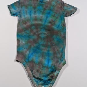 Puede incluir: Un body de beb&eacute; de manga corta con un estampado tie-dye en tonos azules, turquesas y marrones. El body tiene un cuello redondo y corchetes en la parte inferior. La tela parece suave y c&oacute;moda.
