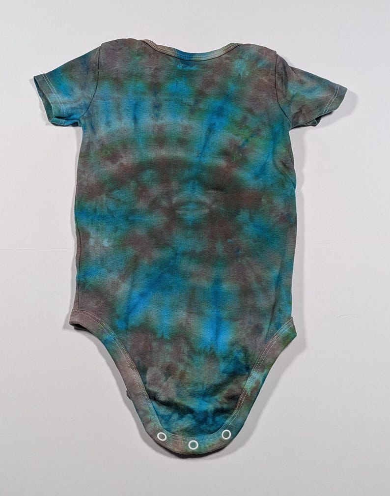 Puede incluir: Un body de beb&eacute; de manga corta con un estampado tie-dye en tonos azules, turquesas y marrones. El body tiene un cuello redondo y corchetes en la parte inferior. La tela parece suave y c&oacute;moda.