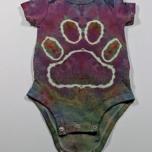 May include: Tie-dye baby onesie in shades of purple, blue, and green. The onesie features a large white paw print design on the front. The garment has short sleeves and snaps at the bottom for easy changes.