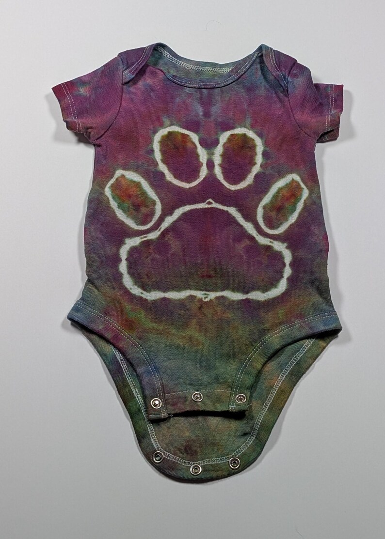 May include: Tie-dye baby onesie in shades of purple, blue, and green. The onesie features a large white paw print design on the front. The garment has short sleeves and snaps at the bottom for easy changes.