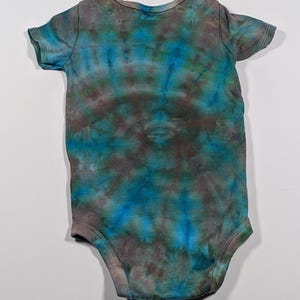 Puede incluir: Un body de beb&eacute; te&ntilde;ido con tinte en tonos azules, turquesas y marrones. La prenda de manga corta tiene un cuello redondo y corchetes en la parte inferior para facilitar los cambios. La tela tiene un patr&oacute;n arremolinado.