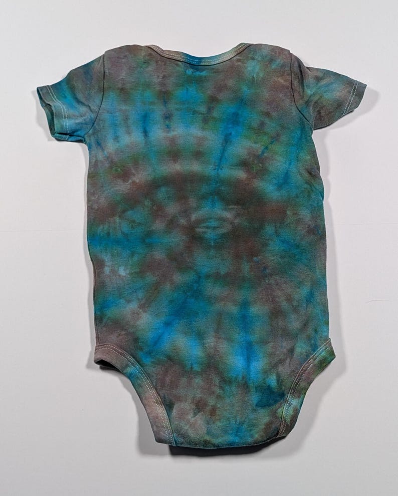 Puede incluir: Un body de beb&eacute; te&ntilde;ido con tinte en tonos azules, turquesas y marrones. La prenda de manga corta tiene un cuello redondo y corchetes en la parte inferior para facilitar los cambios. La tela tiene un patr&oacute;n arremolinado.