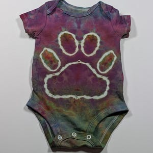 May include: A tie-dye baby onesie in shades of purple, green, and blue, featuring a large white paw print design. The onesie has short sleeves and three snap closures at the bottom. The fabric has a mottled, watercolor effect.