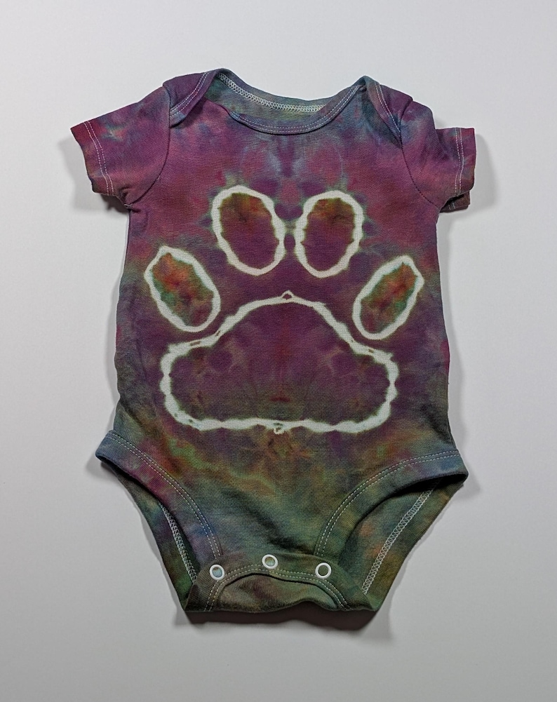May include: A tie-dye baby onesie in shades of purple, green, and blue, featuring a large white paw print design. The onesie has short sleeves and three snap closures at the bottom. The fabric has a mottled, watercolor effect.