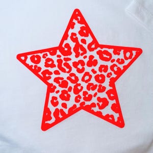 May include: A white t-shirt with a large red star design. The star is outlined in red and filled with a red leopard print pattern. The shirt has a simple, classic design.