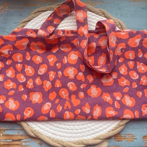 Könnte beinhalten: Rechteckige Tragetasche mit einem orangefarbenen Leopardenmuster auf lila Grund. Die Tasche hat passende Griffe und liegt auf einer weißen und beigefarbenen gewebten Matte.