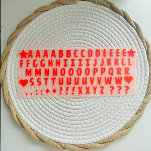 Op de afbeelding: Een transparant vel met rode alfabetletters en symbolen, waaronder sterren en harten, wordt weergegeven op een wit en beige geweven placemat. De letters zijn in een schreefloze lettertype. De placemat heeft een cirkelvorm.