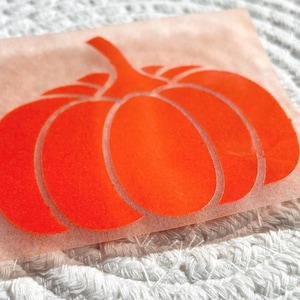 Op de afbeelding: Een oranje pompoenvormig warmteoverdracht vinylontwerp op een perzik kleurige achtergrond. De pompoen heeft een steel en bestaat uit verschillende afgeronde secties. Het ontwerp is geplaatst op een wit, getextureerd oppervlak.