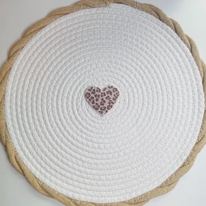 Op de afbeelding: Een ronde, witte geweven placemat met een bruine touwrand. Een kleine hartvormige applicatie met een luipaardprint is gecentreerd op de mat. De placemat is ontworpen met concentrische cirkels.