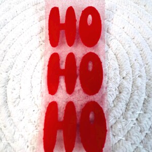 Könnte beinhalten: Ein rechteckiger, transparenter Aufkleber mit dem Wort „HO“ dreimal in fetten, roten Buchstaben. Der Aufkleber befindet sich vor einem weißen, strukturierten Hintergrund und erzeugt ein festliches Urlaubsgefühl.