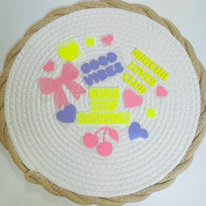 Könnte beinhalten: Ein runder, weißer Platzdeckchen mit gewebter Textur, umrandet von einem braunen Seil. Verziert mit farbenfrohen, durchscheinenden Aufklebern, darunter Herzen, Sterne, eine Schleife, Kirschen und Sätze wie "Good Vibes" und "Matcha Lover Club."