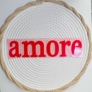 Op de afbeelding: Een ronde, wit geweven placemat met een beige touwrand. Het woord "amore" staat in felrode letters over het midden van de placemat. De placemat is ontworpen voor decoratief gebruik.