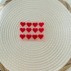 Puede incluir: Una hoja de pegatinas rojas en forma de corazón sobre un fondo rosa claro. Las pegatinas están dispuestas en un patrón de cuadrícula. Las pegatinas están sobre un salvamanteles blanco tejido.