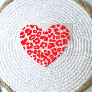 Peut inclure: Un dessous de plat rond blanc tressé avec un motif en forme de cœur. Le cœur est rempli d'un imprimé léopard rouge. Le bord du dessous de plat a une bordure en corde de couleur naturelle.