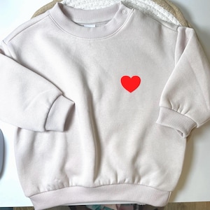 Puede incluir: Sudadera beige claro con un corazón rojo en la parte delantera. La parte superior de manga larga tiene puños acanalados y cuello redondo con un solo botón en el hombro. La sudadera está hecha de un material suave.