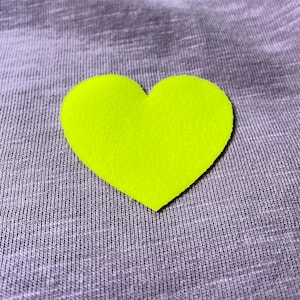 May include: A bright neon yellow heart-shaped felt applique. The heart is centered on a gray, textured fabric background. The felt has a slightly rough texture.