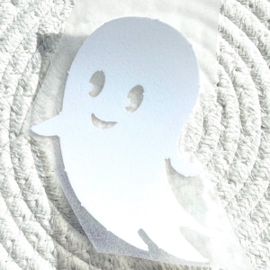 Peut inclure: Autocollant blanc en forme de fantôme souriant, avec deux découpes pour les yeux et une bouche incurvée. L'autocollant est sur un support transparent, prêt à être appliqué. Le fond est une surface texturée blanc cassé.