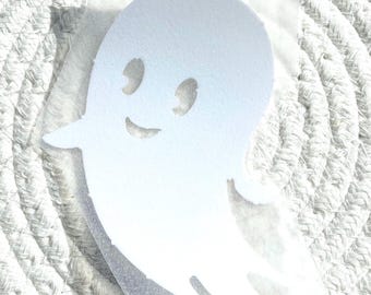 May include: A white ghost decal featuring a simple cartoon design with two oval eyes and a curved mouth. The ghost shape is cut out from a matte white material and is attached to a clear, rectangular adhesive backing sheet for easy application.