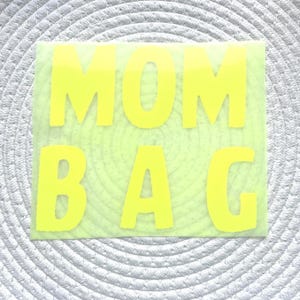 Könnte beinhalten: Ein neongelbes Rechteck mit den Worten "MOM BAG" in fetten Großbuchstaben. Der Text ist mittig auf dem Rechteck platziert, das sich vor einem strukturierten, hellgrauen Hintergrund befindet. Das Design ist schlicht und auffällig.